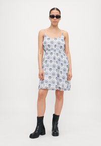 Rochie albă cu model paisley albastru, bretele subțiri și un corset ajustat. Asortată cu cizme negre supradimensionate și ochelari de soare.