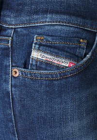 Jeans de mezclilla azul oscuro con un remache de cobre y una pequeña etiqueta que dice "Diesel Industry" en el bolsillo trasero. Textura suave.