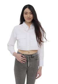 Camicia bianca cropped con bottoni, dotata di due tasche frontali, abbinata a jeans grigi a vita alta. Tessuto liscio, design strutturato e vestibilità su misura.
