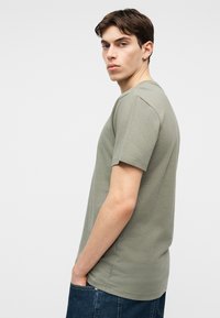 Νεαρός άνδρας με πράσινο κοντομάνικο μπλουζάκι και μπλε τζιν στέκεται πλαγίως με το ένα χέρι στην τσέπη, κοιτάζοντας ελαφρώς προς την κάμερα.