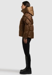 Braune Steppjacke mit hohem Kragen und Kapuze, mit gestepptem Muster und Seitentaschen, kombiniert mit schwarzen Leggings und kniehohen Stiefeln.