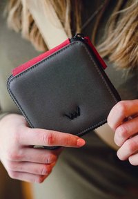 Gray fabric wallet with red zipper pull, rectangular shape, textured surface, and a black logo on the front. Held in a hand.