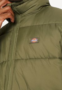 Kurtka puchowa oliwkowa z wysokim kołnierzem, wykonana z teksturowanej tkaniny, z wyraźnym przeszyciem i małą naszywką z logo na lewej piersi.