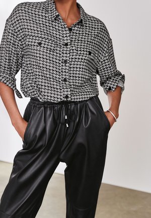 Personne portant une chemise à boutons noir et blanc à motif pied-de-poule avec manches retroussées et un pantalon en cuir noir avec cordon, mains dans les poches.