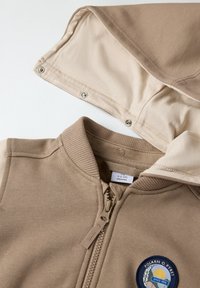 Beige zip-up sweatshirt med ribbet krave, blødt stof og et cirkulært logo-patch med en sol og tekst. Hætten har trykknapper.