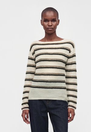 Femme aux cheveux courts portant un pull crème avec des rayures horizontales noires et marron et un jean bleu foncé, debout devant un fond blanc.