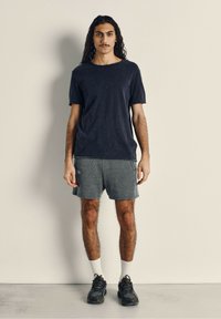 Camiseta de manga corta azul marino, pantalones cortos deportivos grises, calcetines blancos y zapatillas negras. El modelo tiene el cabello largo y está de pie contra un fondo neutro.