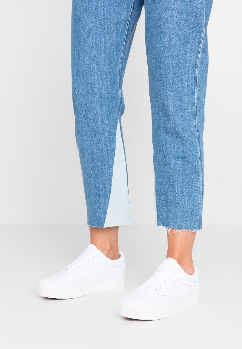 Baskets blanches en toile avec semelles plates, détails du logo et lacets blancs, portées avec un jean denim bleu raccourci présentant un panneau bleu clair.