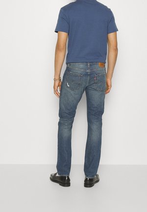Mand iført blå polo shirt, falmede blå jeans med baglommer og sorte pæne sko, stående med ryggen til mod en ensfarvet baggrund.