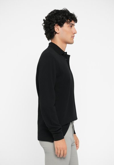Cămașă polo neagră cu mânecă lungă și guler, având un croi drept și o textură subtilă, purtată cu pantaloni gri deschis, vedere laterală.