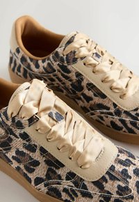 Paire de baskets en tissu imprimé léopard, accents en cuir beige, larges lacets en satin beige, et semelles en caoutchouc marron sur fond blanc.