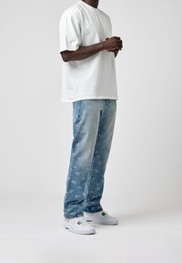 Man med en vit oversized t-shirt, ljusblå mönstrade jeans och vita sneakers med gröna accenter som står mot en enkel bakgrund.