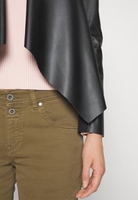 Veste en simili cuir noir avec ourlet asymétrique, associée à un haut à côtes rose pastel et un pantalon vert olive avec deux fermetures à boutons.