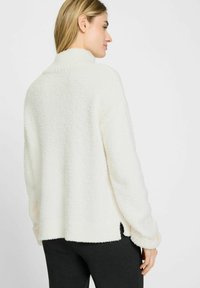 Weißer, flauschiger Pullover mit hohem Kragen, angeschnittenen Schultern und seitlichen Schlitzen. Er zeigt eine strukturierte Oberfläche sowie gerippte Abschlüsse an den Ärmeln und am Saum.