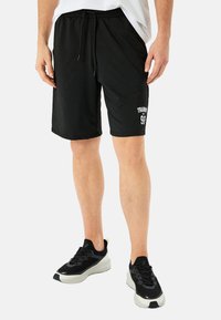 Sorte træningsshorts med elastisk talje og snøre. Funktioner tekst "TRAINING" og nummeret "98." Bæres med sorte træningssko.