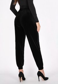 Zwarte fluwelen joggingbroek met een geplooid tailleband en afgeknipte enkels, gecombineerd met zwarte hoge hakken, bekeken vanuit de achterkant.