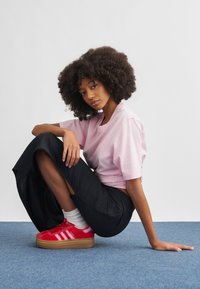 Jovem com cabelo encaracolado sentada de pernas cruzadas num tapete azul, vestindo camisa rosa, calças pretas, meias brancas e ténis vermelhos.