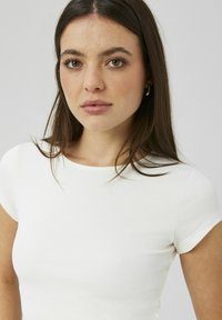 Haut blanc à manches courtes côtelé avec un col rond, présentant une coupe ajustée et une texture lisse. L'arrière-plan est d'une couleur claire neutre.