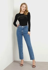 Fekete hosszú ujjú body "Guess 1981" logóval, magas derekú kék egyenes szárú farmerrel és fekete magassarkú szandállal párosítva.