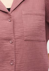 Μωβ πουκάμισο με υφή και κουμπιά εμπρός, με τσέπη στο στήθος, φορεμένο πάνω από μαύρη μπλούζα, εμφανές το γιακά και τρία κουμπιά.