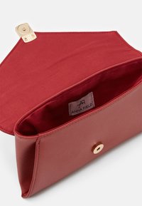 Borsetta a tracolla sintetica rossa con una superficie liscia. Presenta una chiusura a scatto dorata e una fodera in tessuto. Etichetta di marca visibile all'interno.