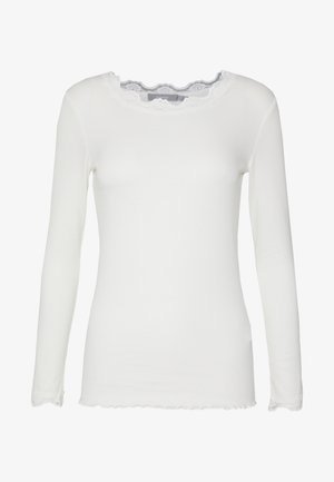 Bela dolga majica z dolgimi rokavi, izdelana iz rebrastega materiala, s čipkastim ovratnikom in robom z valovnim vzorcem. Ima prilegajočo se zasnovo s preprosto teksturo.