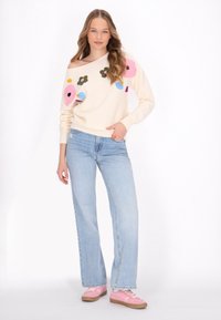 Crème trui met oversized bloemenborduursel in roze, blauw en groen. Gecombineerd met lichtblauwe jeans en roze sneakers, met een relaxed fit.