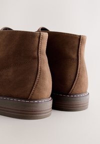 Braune Wildleder-Stiefeletten mit glatter Textur, rundem Zeh und mehrschichtiger Holzsohle. Sie verfügen über saubere Nähte und einen gepolsterten Kragen.