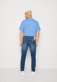 Jeans de denim con un lavado azul medio, con bolsillos traseros, costuras visibles y una etiqueta. Combinados con una camiseta azul clara y zapatillas blancas.