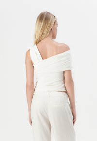 Blusa blanca sin mangas con tejido drapeado, que presenta un diseño torcido, combinada con pantalones de color beige claro y una textura suave.
