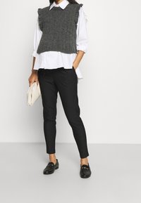 Chaleco de punto gris con hombros con volantes sobre una blusa blanca, combinado con pantalones negros de rayas y mocasines negros. Sostiene un bolso de mano color crema.