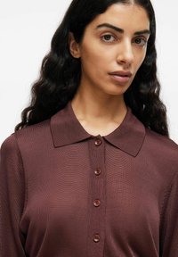 Donna con lunghi capelli ricci e scuri che indossa una camicia bordeaux con colletto, dotata di bottoni e texture, su uno sfondo bianco uniforme.