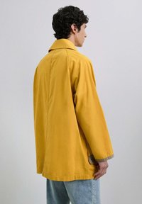 Chaqueta de algodón amarilla con un corte relajado, diseño oversize, solapa ancha y acentos beige contrastantes cerca de los puños.