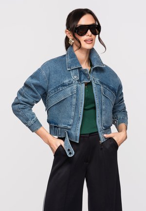 Žena v nadměrné džínové bundě, tmavě zeleném topu, černých širokých kalhotách, velkých slunečních brýlích a zlatých výrazných náušnicích, pózující s rukama v kapsách.
