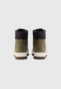 Une paire de bottes en daim vert olive avec des cols rembourrés noirs, des semelles intermédiaires blanches et des semelles extérieures en caoutchouc marron, présentées de l'arrière sur un fond blanc.