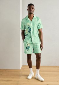 Grøn skjorte med korte ærmer og knaplukning samt matchende shorts, prydet med blomsterbroderi. Fremstillet af letvægtsstof med en blød, tekstureret finish.