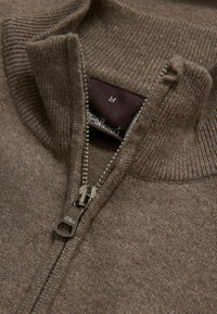 Bruine zip-up sweater gemaakt van zacht materiaal, met een ribgebreide kraag, ritssluiting en een label met maat "M" aan de binnenkant.