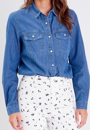 Femme portant une chemise en denim bleu à manches longues avec boutons et un pantalon blanc à petit imprimé floral noir.
