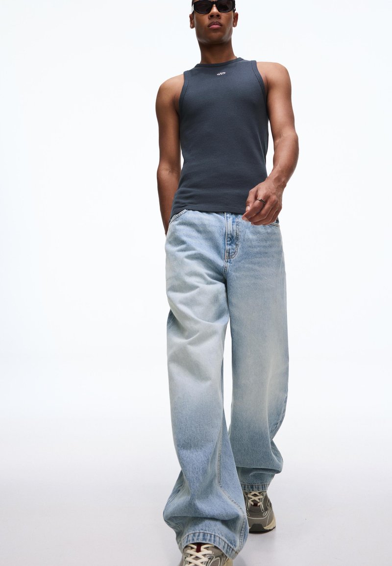 Grijze tanktop met smalle bandjes, lichtblauwe wijde jeans en sneakers met patroon. Model draagt een donkere zonnebril en staat tegen een witte achtergrond.