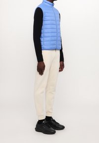 Světle modrá péřová vesta s stojáčkem, nošená přes černou dlouhou košili, v kombinaci s krémovými tepláky a černými teniskami.