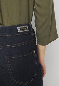 Mörkblå denimjeans med en figurnära design. Har silverfärgad metall, kontrasterande sömmar och en logotyp på midjebandet.