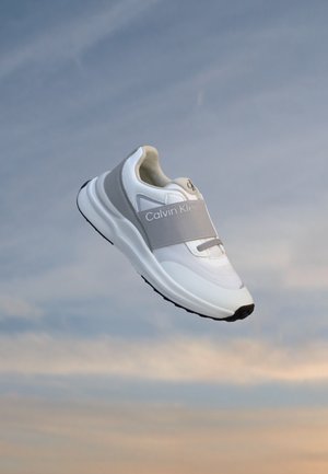 Hvid atletiksko med grå rem mærket Calvin Klein, der svæver mod en blød himmelbaggrund ved solnedgang.