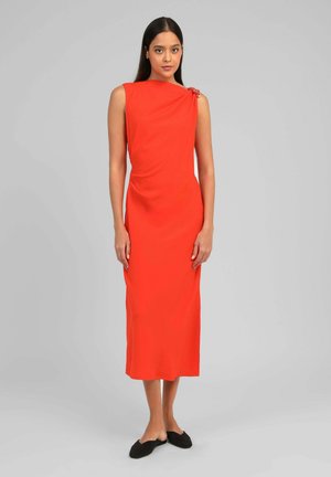 Femme debout portant une robe midi sans manches de couleur orange vif avec un détail froncé et des chaussures plates noires, sur fond gris.