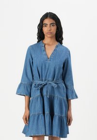 Denim ruha V-nyakkivágással és háromnegyedes harangujjakkal. Réteges fodrokkal, megkötővel ellátott, karcsúsított derékkal, világoskék színben.