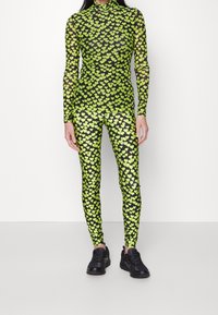 Schwarzes Mesh-Bodysuit und Leggings mit neongrünem Herzmuster. Lange Ärmel, hoher Kragen und kombiniert mit schwarzen Sneakers.