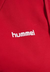 Rode stof met witte "hummel" logo tekst gedrukt nabij zichtbare trekkoord aan de rechterkant van het kledingstuk.