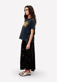 Frau mit langen, dunklen Haaren, die ein schwarzes Grafik-T-Shirt, einen schwarzen Spitzen-Maxikleid und metallic-silberne Slipper trägt, seitlich vor weißem Hintergrund stehend.