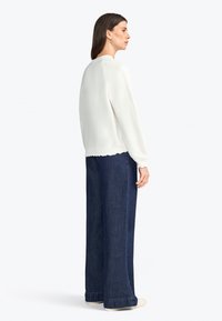 Top bianco a maniche lunghe con orlo scallop, abbinato a jeans in denim scuro a gamba larga. Modella vista di lato su uno sfondo neutro.