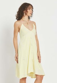 Mujer con cabello rizado lleva un vestido sin mangas de color amarillo pálido con tirantes finos y sutiles detalles decorativos, de pie frente a un fondo liso.
