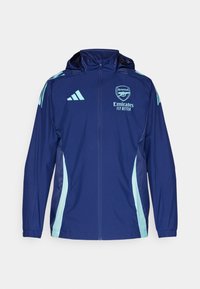 Giacca windbreaker navy con accenti azzurri, zip completa, cappuccio e logo Adidas. Presenta il branding dell'Arsenal sul petto. Materiale leggero.